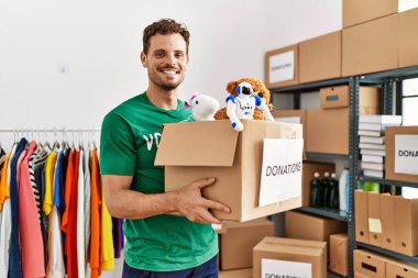 Gönüllü üniformalı genç İspanyol bir adam yardım merkezinde oyuncaklarla dolu bir kutu tutuyor.