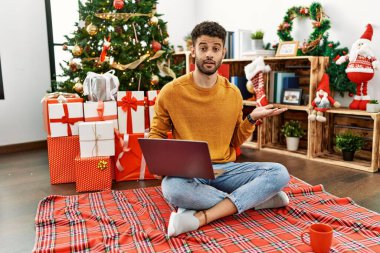 Arap genç adam dizüstü bilgisayar kullanıyor Noel ağacının yanında oturuyor avuç içi açık bir şekilde fotokopi alanını gösteriyor, gülümseyerek mutlu bir gülümseme sergiliyor. 