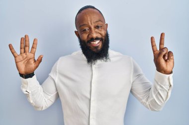 Afro-Amerikalı adam mavi arka planda duruyor ve yedinci parmaklarını gösterip işaret ederken kendinden emin ve mutlu gülümsüyor.. 