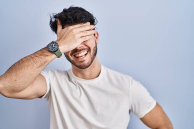 Hispanic man with beard standing over white background smiling and laughing with hand on face covering eyes for surprise. blind concept. 
