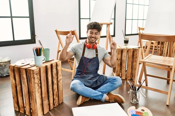 Young Hispanic Man Sitting Art Studio Gesturing Finger Crossed Smiling — Stockfoto