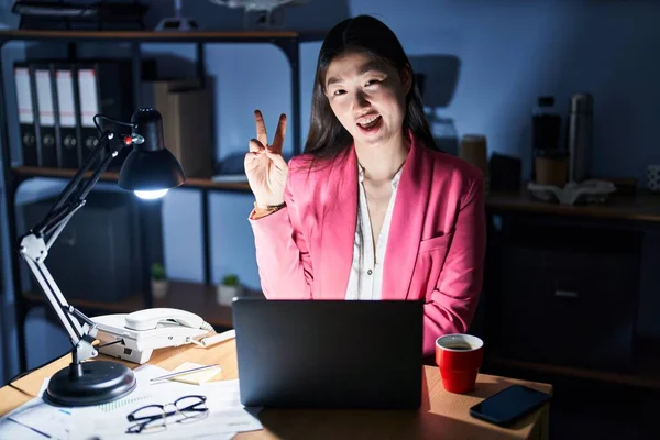 Chinese young woman working at the office at night smiling with happy face winking at the camera doing victory sign. number two.