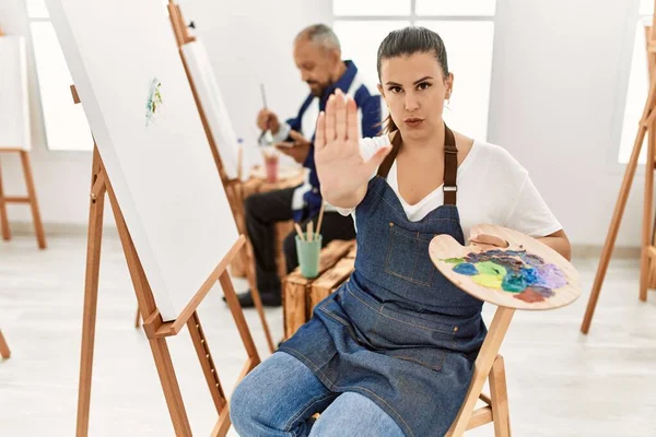 Young artist woman at art studio with open hand doing stop sign with serious and confident expression, defense gesture