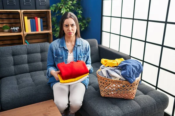 Hispanic woman holding folded laundry after ironing skeptic and nervous, frowning upset because of problem. negative person.