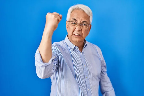 Hispanic senior man wearing glasses angry and mad raising fist frustrated and furious while shouting with anger. rage and aggressive concept.