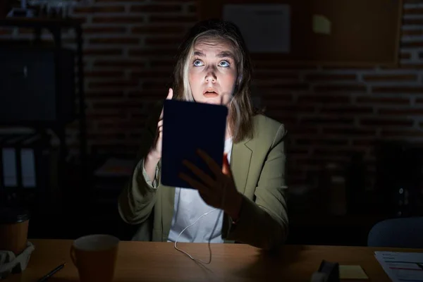 Blonde caucasian woman working at the office at night pointing up looking sad and upset, indicating direction with fingers, unhappy and depressed.