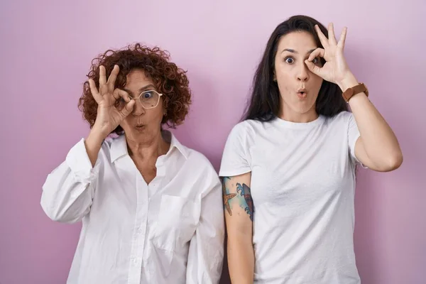 ピンクの背景にカジュアルな白いTシャツを着たヒスパニック系の母親と娘は 驚きの顔でショックを受けたOkジェスチャーを行うと 目の指を見て 信じられない表現 — ストック写真