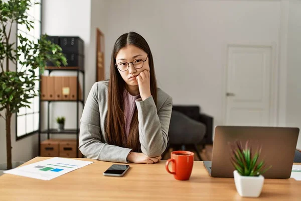 Young chinese business worker wearing business style sitting on desk at office thinking looking tired and bored with depression problems with crossed arms.