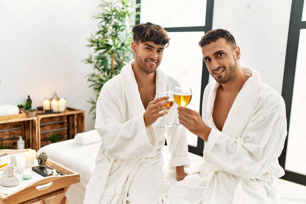 Two hispanic men couple toasting with champagne sitting on massage table at beauty center