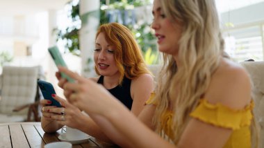 Two women using smartphones and drinking coffee sitting on table at home terrace
