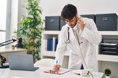 Young hispanic man wearing doctor uniform talking on telephone writing on document at clinic