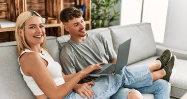 Evdeki kanepede oturup dizüstü bilgisayar kullanarak gülümseyen beyaz bir çift..