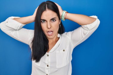 Mavi arka planda dikilen Latin kökenli genç kadın deli ve elleri başının üstünde korkmuş, korkmuş ve ağzı açık şoktan şaşırmış. 
