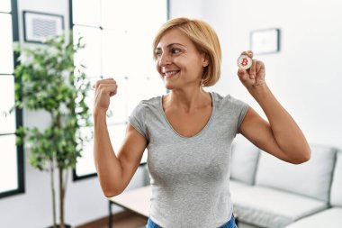 Orta yaşlı sarışın kadın elinde sanal para tutuyordu. Bitcoin parmağıyla yan tarafı gösteriyordu. Mutlulukla gülümsüyordu. 