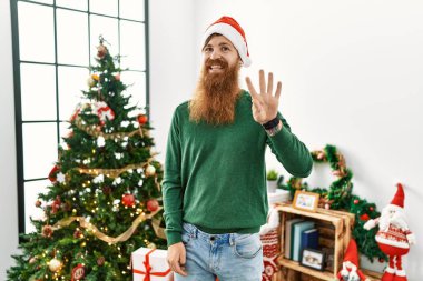 Redhead man with long beard wearing christmas hat by christmas tree showing and pointing up with fingers number four while smiling confident and happy. 