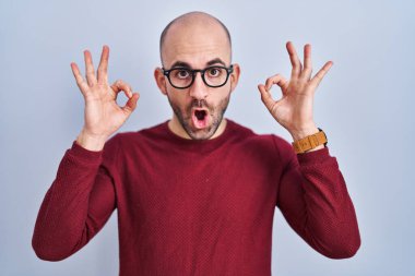 Beyaz arka planda duran sakallı, gözlüklü, şaşırmış ve şok olmuş genç bir adam parmaklarıyla onay sembolü yapıyor. çılgın ifade 