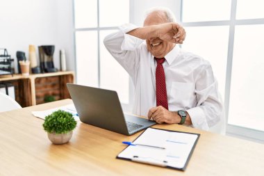 Senior man working at the office using computer laptop covering eyes with arm smiling cheerful and funny. blind concept. 