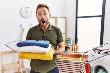 Middle age man holding folded laundry after ironing afraid and shocked with surprise and amazed expression, fear and excited face. 