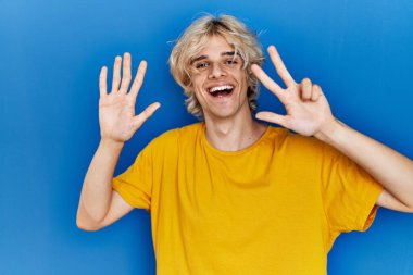 Genç modern adam mavi arka planda duruyor. Sekiz numaralı parmaklarıyla işaret ediyor ve gülümsüyor. Kendinden emin ve mutlu.. 