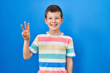 Mavi arka planda duran genç beyaz çocuk üç numarayı gösterip işaret ederken kendinden emin ve mutlu gülümsüyor.. 