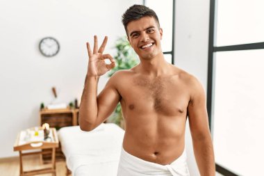 İspanyol asıllı genç adam spa merkezinde üstsüz durup gülümsüyor ve el ve parmaklarıyla 