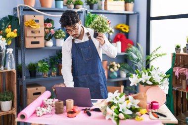 Young hispanic man florist using laptop holding plant at florist shop