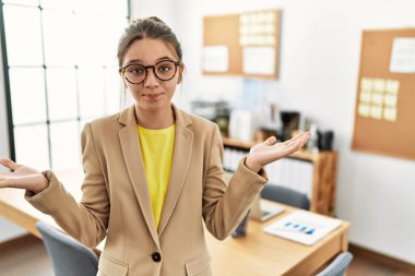 Young brunette teenager wearing business style at office clueless and confused expression with arms and hands raised. doubt concept. 