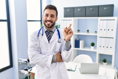 Yakışıklı, doktor üniforması giyen ve klinikte steteskop takan yakışıklı bir adam mutlu bir şekilde elini ve parmağını yan tarafına uzatarak gülümsüyor. 