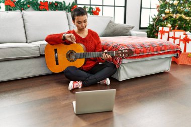 Kısa saçlı İspanyol bir kadın, online öğretmenden klasik gitar öğreniyor. Kızgın suratlı, negatif işaretli, baş parmakları aşağıda, reddedilme kavramından hoşlanmadığını gösteriyor. 