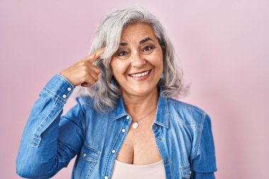 Middle age woman with grey hair standing over pink background smiling pointing to head with one finger, great idea or thought, good memory 