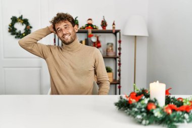 Masada sakallı genç yakışıklı adam Noel süslemeleri ile oturuyor. Kendine güvenen, el hareketleriyle saçlara dokunan, çekici ve modaya uygun poz veren. 