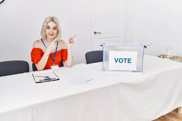 Young caucasian woman at political election sitting by ballot smiling and looking at the camera pointing with two hands and fingers to the side.