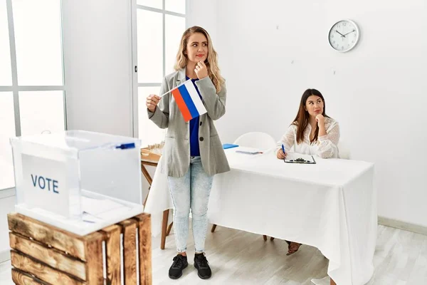 Young blonde woman at political stand holding russia flag serious face thinking about question with hand on chin, thoughtful about confusing idea