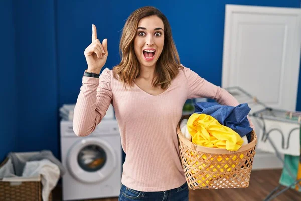 Young woman holding laundry basket pointing finger up with successful idea. exited and happy. number one.