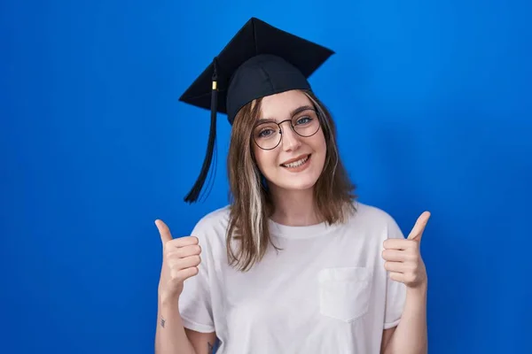 Blonde caucasian woman wearing graduation cap success sign doing positive gesture with hand, thumbs up smiling and happy. cheerful expression and winner gesture.