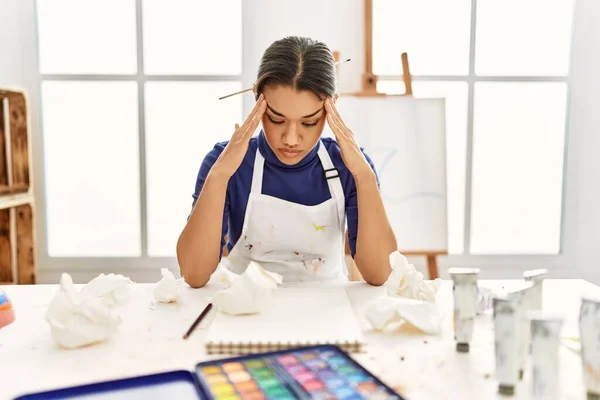 Young latin woman with serious expression sitting on table at art studio