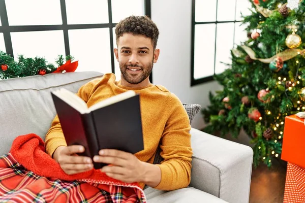 Young arab man reading book lying on the sofa by christmas tree at home.