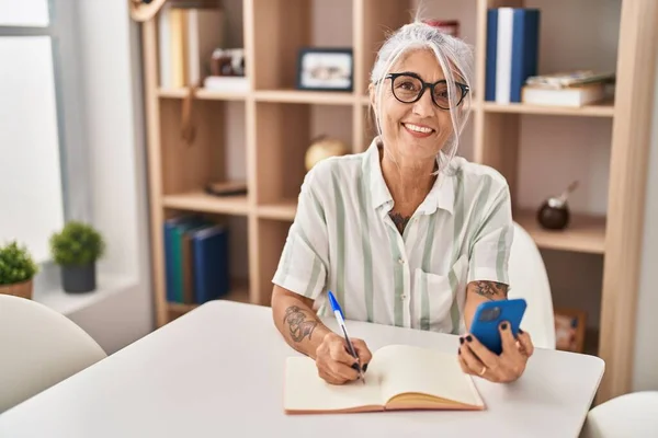 Mujer Pelo Gris Mediana Edad Escribiendo Cuaderno Usando Teléfono Inteligente —  Fotos de Stock