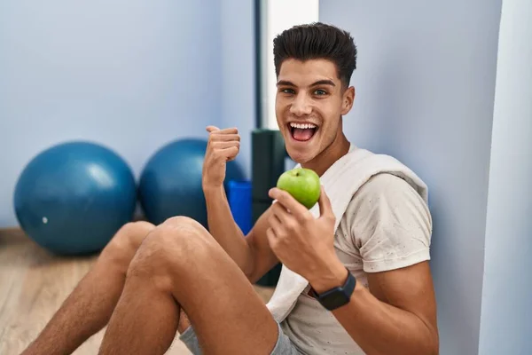 Young Hispanic Man Wearing Sportswear Eating Healthy Apple Pointing Thumb — Stockfoto