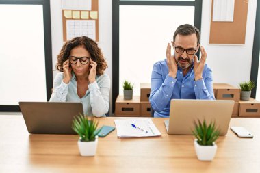 Middle age hispanic woman and man sitting with laptop at the office with hand on head for pain in head because stress. suffering migraine. 