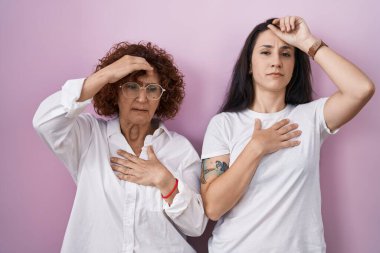 İspanyol anne ve kızı pembe arka plan üzerine gündelik beyaz tişört giyip hastalık ve ateş için alnına dokunuyorlar, grip ve soğuk algınlığı, virüs hastalığı. 