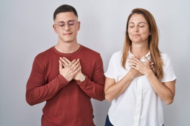Mother and son standing together over isolated background smiling with hands on chest with closed eyes and grateful gesture on face. health concept. 