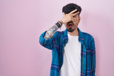 Young hispanic man with beard standing over pink background peeking in shock covering face and eyes with hand, looking through fingers with embarrassed expression. 