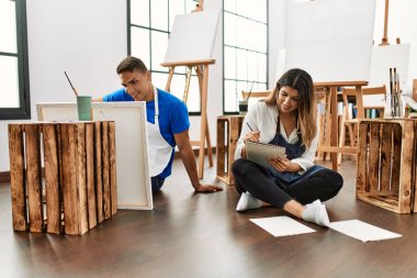 Resim yaparken gülümseyen iki öğrenci sanat okulunda yerde oturuyor..