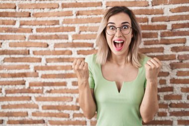 Young beautiful woman standing over bricks wall celebrating surprised and amazed for success with arms raised and open eyes. winner concept. 