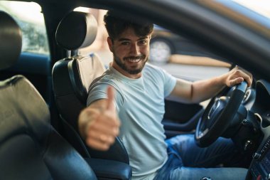 Young hispanic man driving car doing ok gesture with thumb up at street