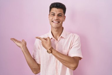Young hispanic man standing over pink background amazed and smiling to the camera while presenting with hand and pointing with finger. 