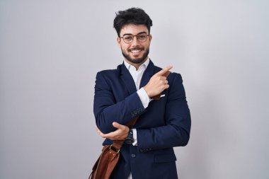 Hispanic man with beard wearing business clothes with a big smile on face, pointing with hand finger to the side looking at the camera. 