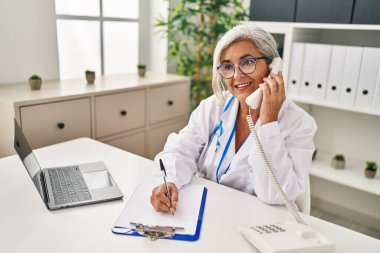 Klinikte doktor üniforması giyen orta yaşlı bir kadın telefonla konuşuyor.