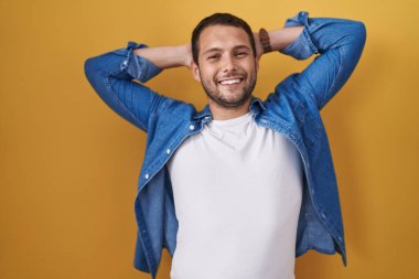 Hispanic man standing over yellow background relaxing and stretching, arms and hands behind head and neck smiling happy 